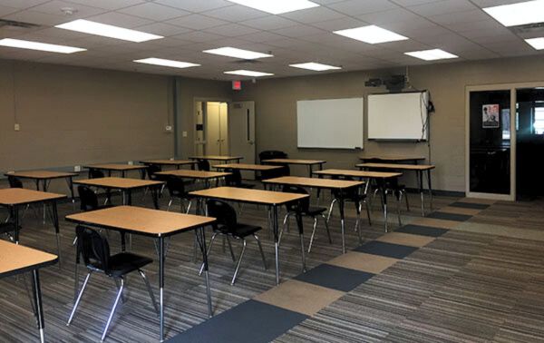  A classroom-style activity room with rows of wooden desks and black chairs, whiteboards mounted on the wall, and overhead fluorescent lighting. The room features a striped carpet floor and an open door leading to another space.