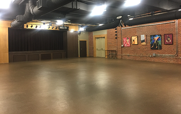 An auditorium with an empty floor space, exposed brick walls, and a small stage with curtains at one end. Overhead lighting and black ceiling ducts are visible, and colorful artwork is displayed on the wall. A set of double doors leads to another area.