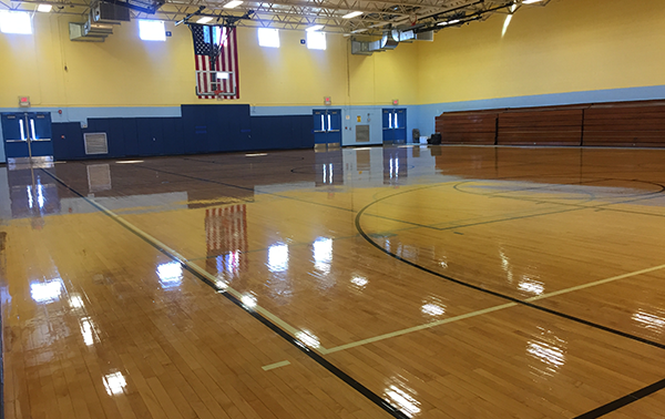 A spacious gymnasium with a polished wooden floor, marked with basketball court lines. The walls are painted yellow and blue, and an American flag hangs at the far end. The gym features bleacher seating on one side and several doors leading to other areas.